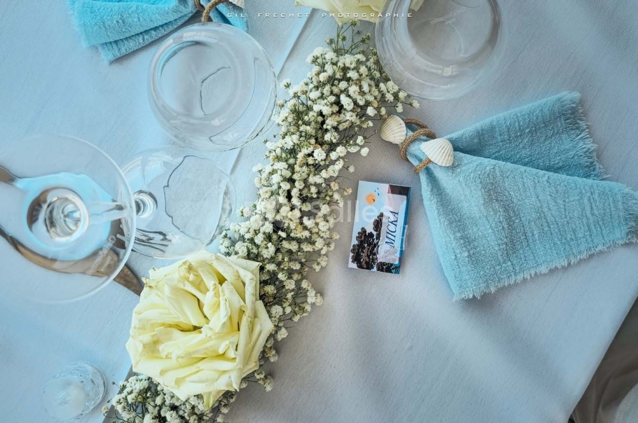 Serviette bleue pliée avec un nœud, rose blanche et gypsophile sur une table dressée avec des verres.