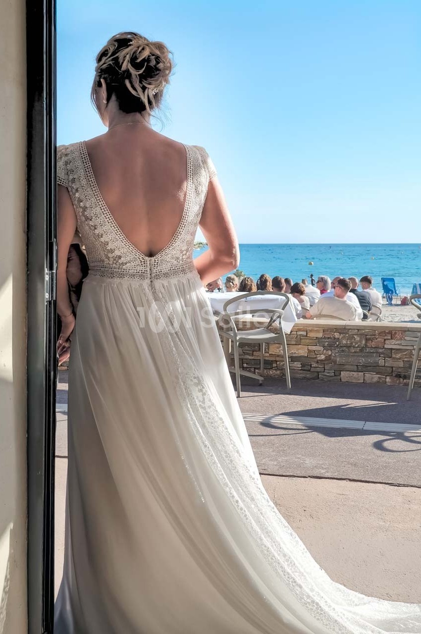 Une femme en robe de mariée dos à la caméra, face à une terrasse en bord de mer par une journée ensoleillée.