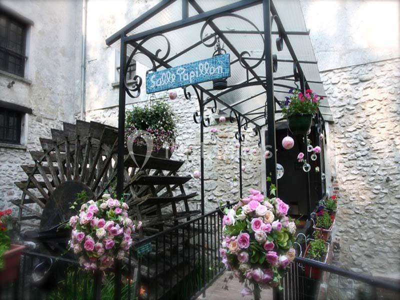 Passage couvert décoré de fleurs roses et blanches, avec une roue à eau en arrière-plan et un panneau suspendu.