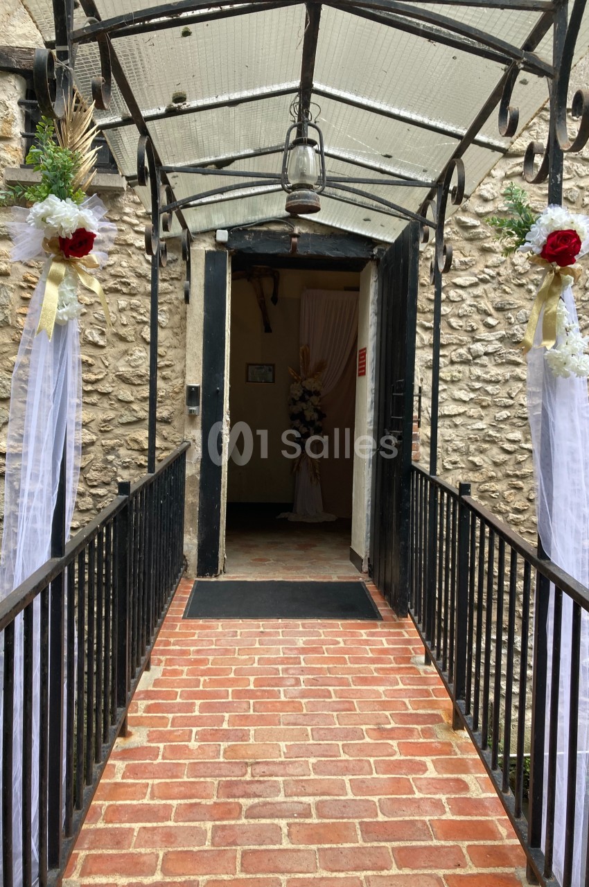 Entrée en briques avec une arche métallique décorée de fleurs blanches et rouges menant à une porte ouverte.