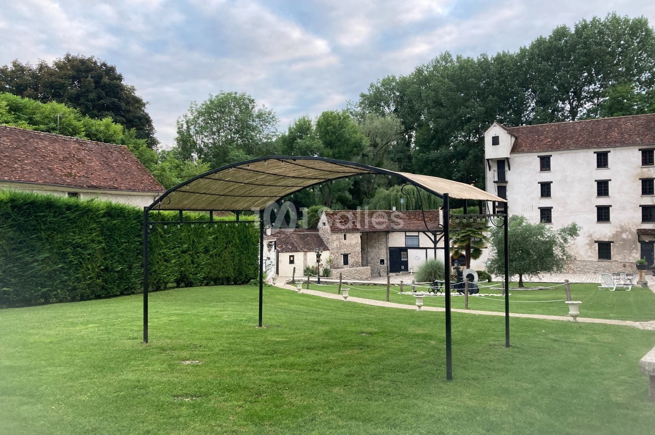 Structure métallique avec toit en toile dans un jardin verdoyant, entouré de bâtiments anciens et d'arbres.