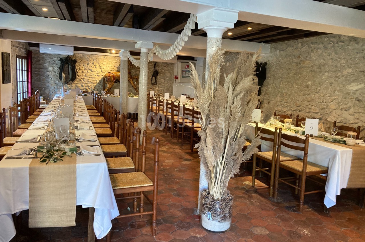 Salle de réception avec tables alignées, nappes blanches, décorations végétales et murs en pierre apparente.