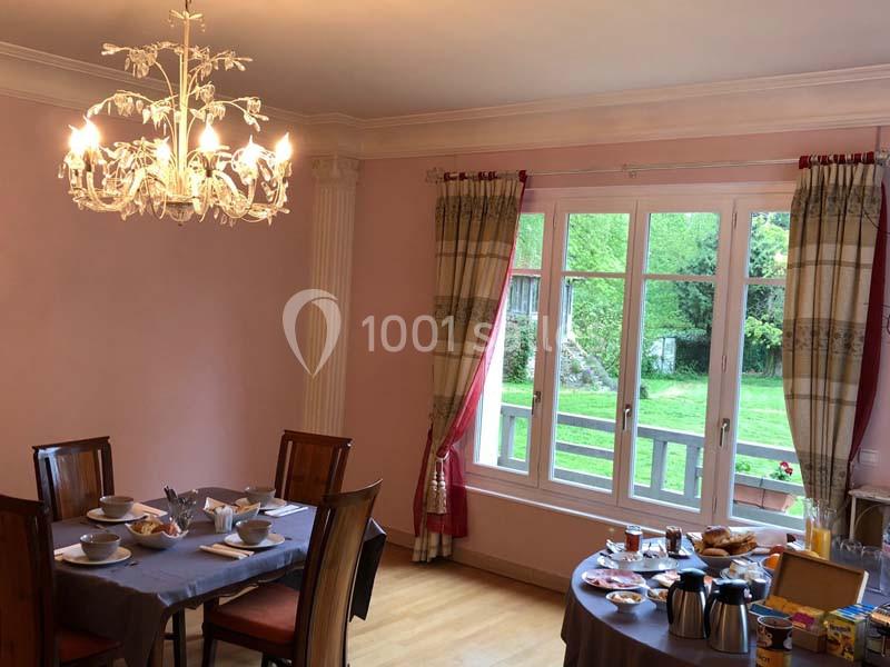 Salle à manger avec tables dressées pour le petit-déjeuner, éclairée par un lustre et donnant sur un jardin verdoyant.