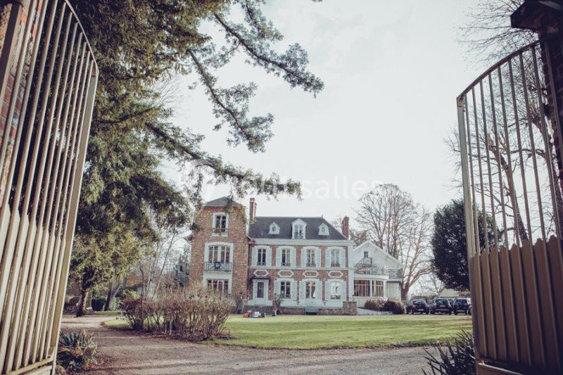 Façade d'une grande maison en briques entourée d'un jardin, vue depuis une entrée avec portail ouvert.