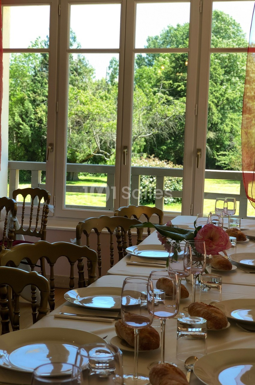 Table dressée avec vaisselle, verres et pain, devant une fenêtre ouverte sur un jardin verdoyant.