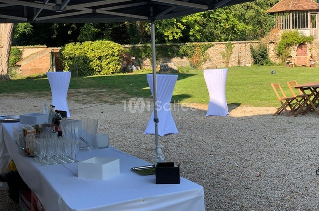 Table dressée sous un barnum avec verres et boissons, dans un jardin avec des tables hautes et un kiosque en arrière-plan.