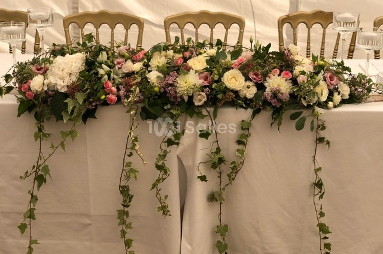 Arrangement floral coloré avec roses, pivoines et feuillage tombant, décorant une table recouverte d'une nappe blanche.