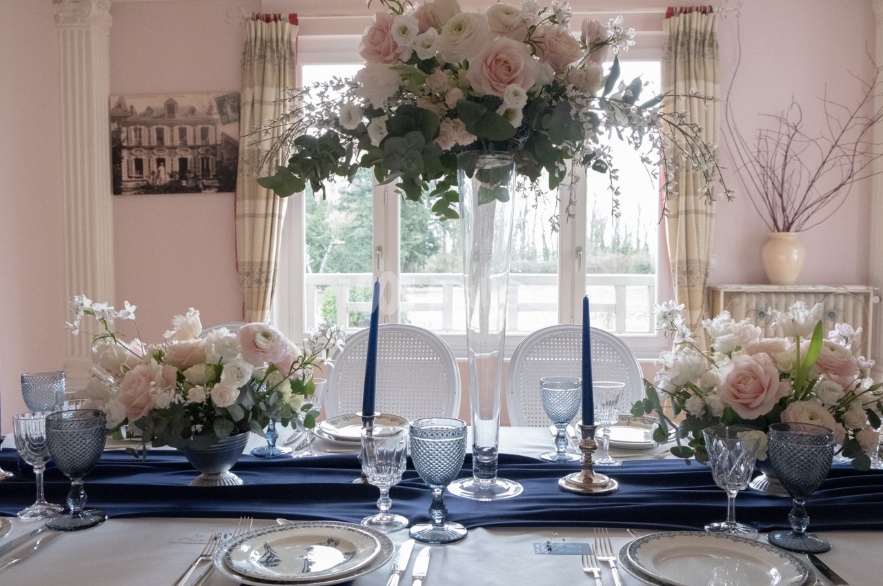 Table élégamment dressée avec nappes bleues, vaisselle raffinée et compositions florales roses et blanches.