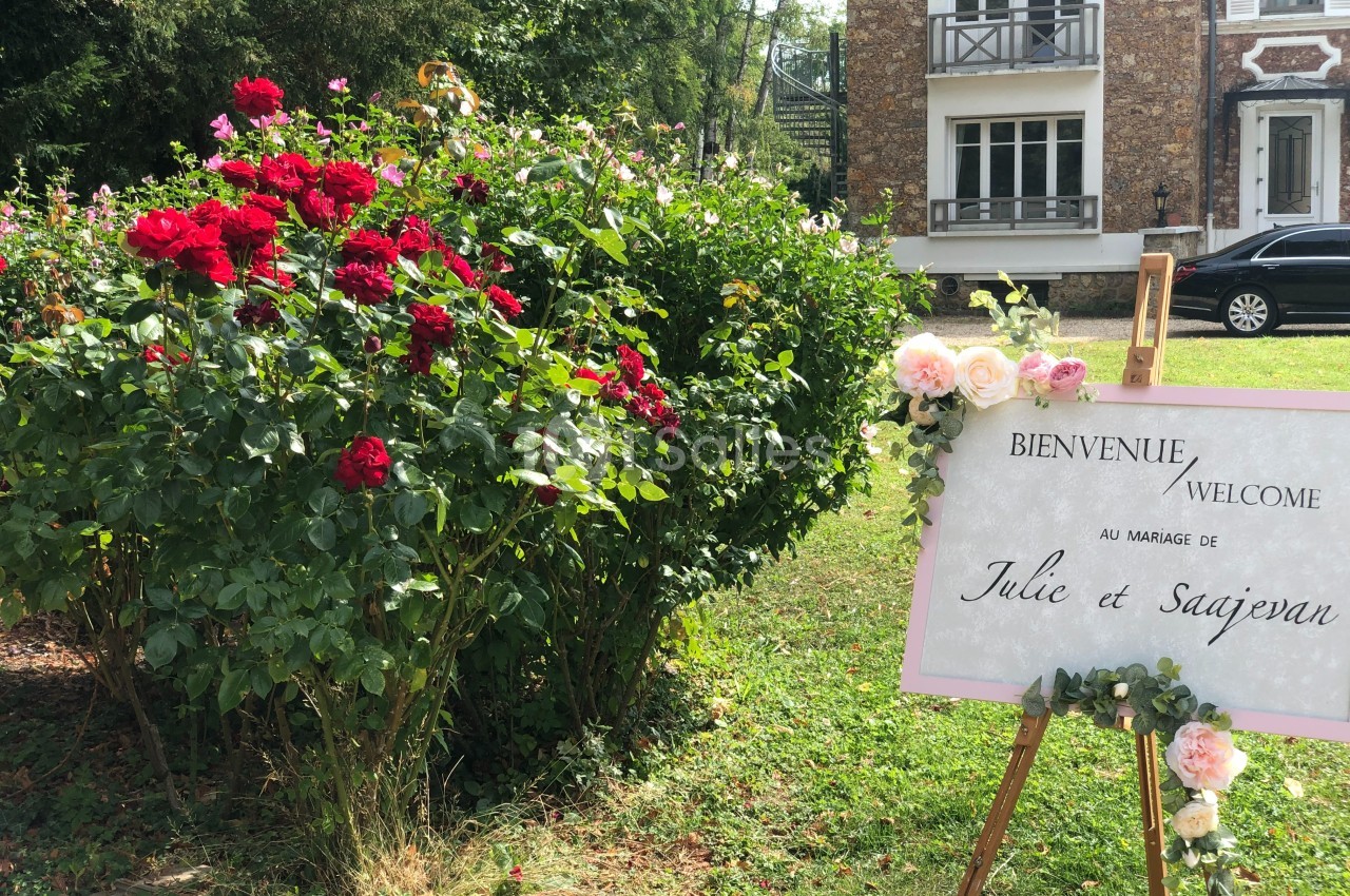 Panneau de bienvenue à un mariage, posé sur un chevalet près d'un massif de roses dans un jardin devant une maison.