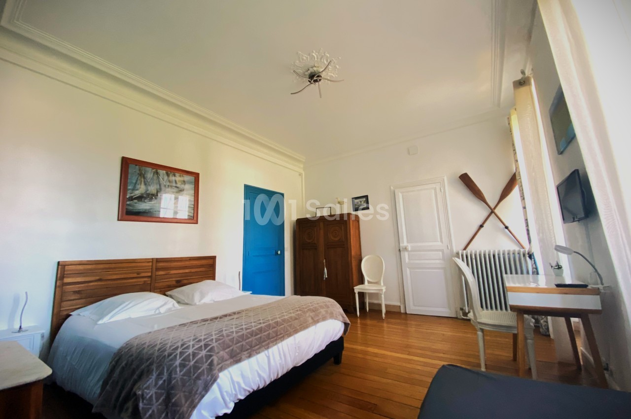 Chambre lumineuse avec lit double, parquet en bois, armoire, décoration murale et fenêtre avec rideaux clairs.