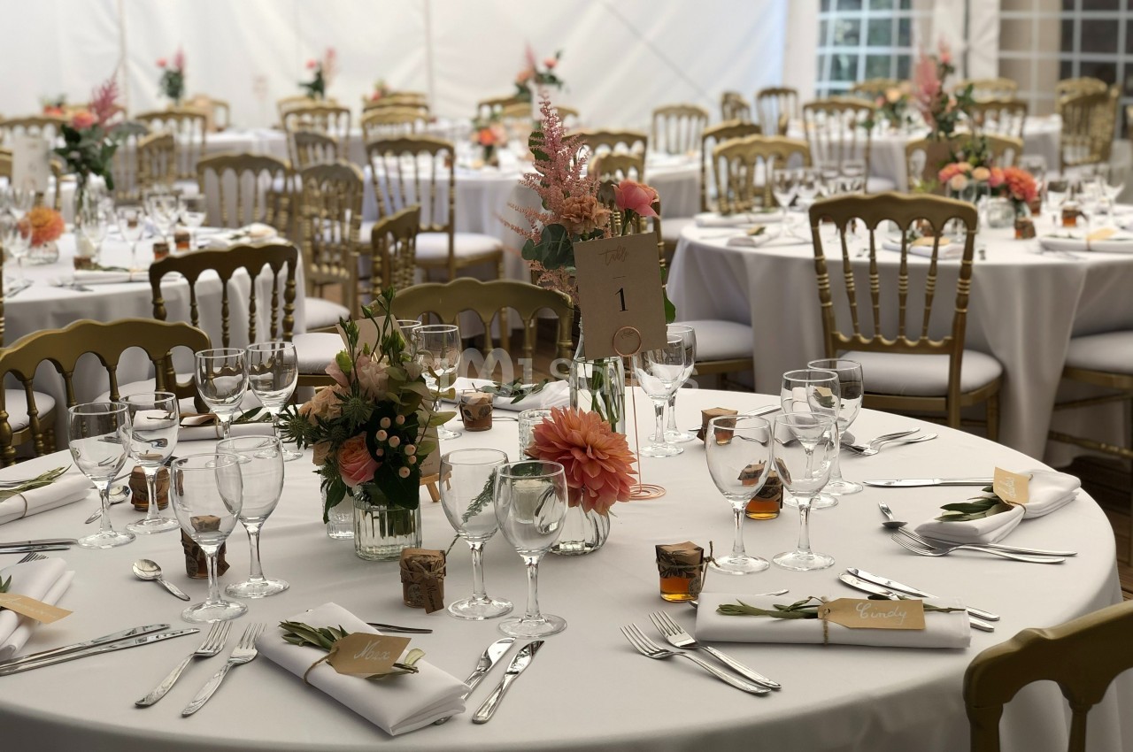 Tables élégamment dressées pour un événement, avec nappes blanches, fleurs, verres et chaises dorées dans une salle…