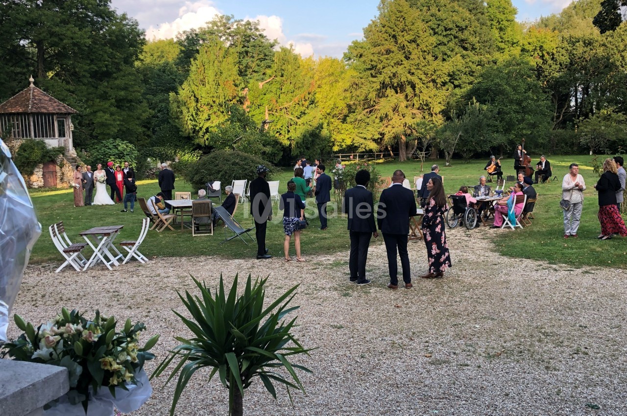 Groupe de personnes rassemblées dans un jardin pour un événement, avec des tables, des chaises et un kiosque en arrière-plan.