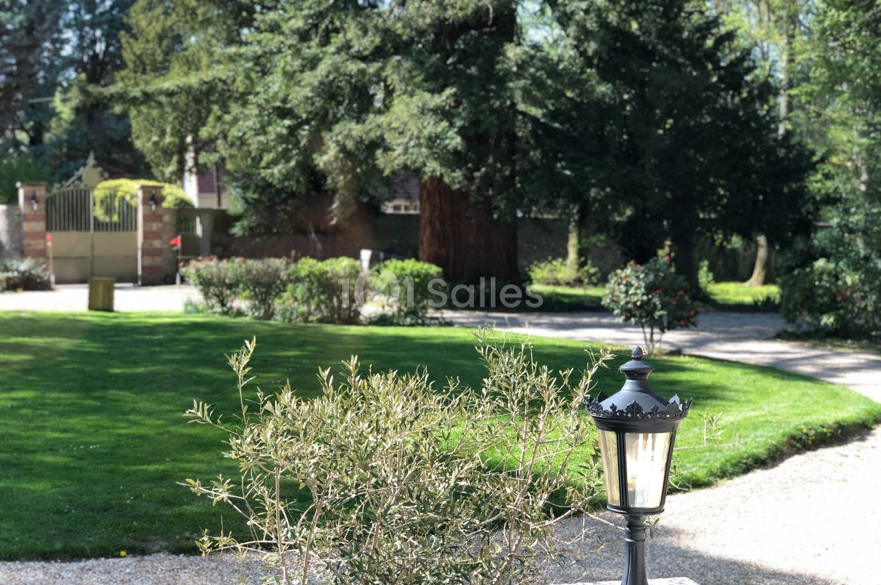 Lanterne en métal noir éclairée, posée sur un muret, devant un jardin verdoyant avec allées gravillonnées et arbres.