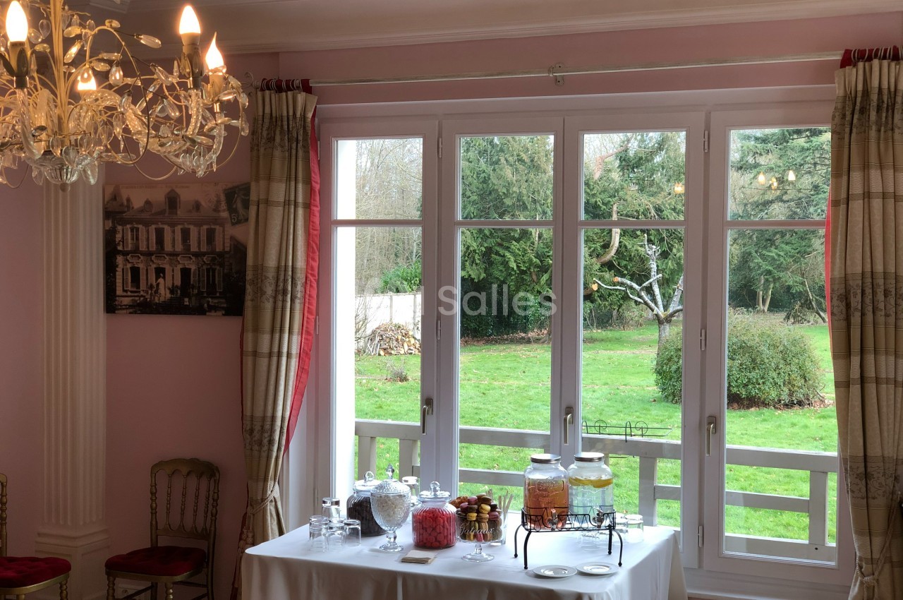 Table dressée avec des boissons et des bonbonnières devant une grande fenêtre donnant sur un jardin verdoyant.