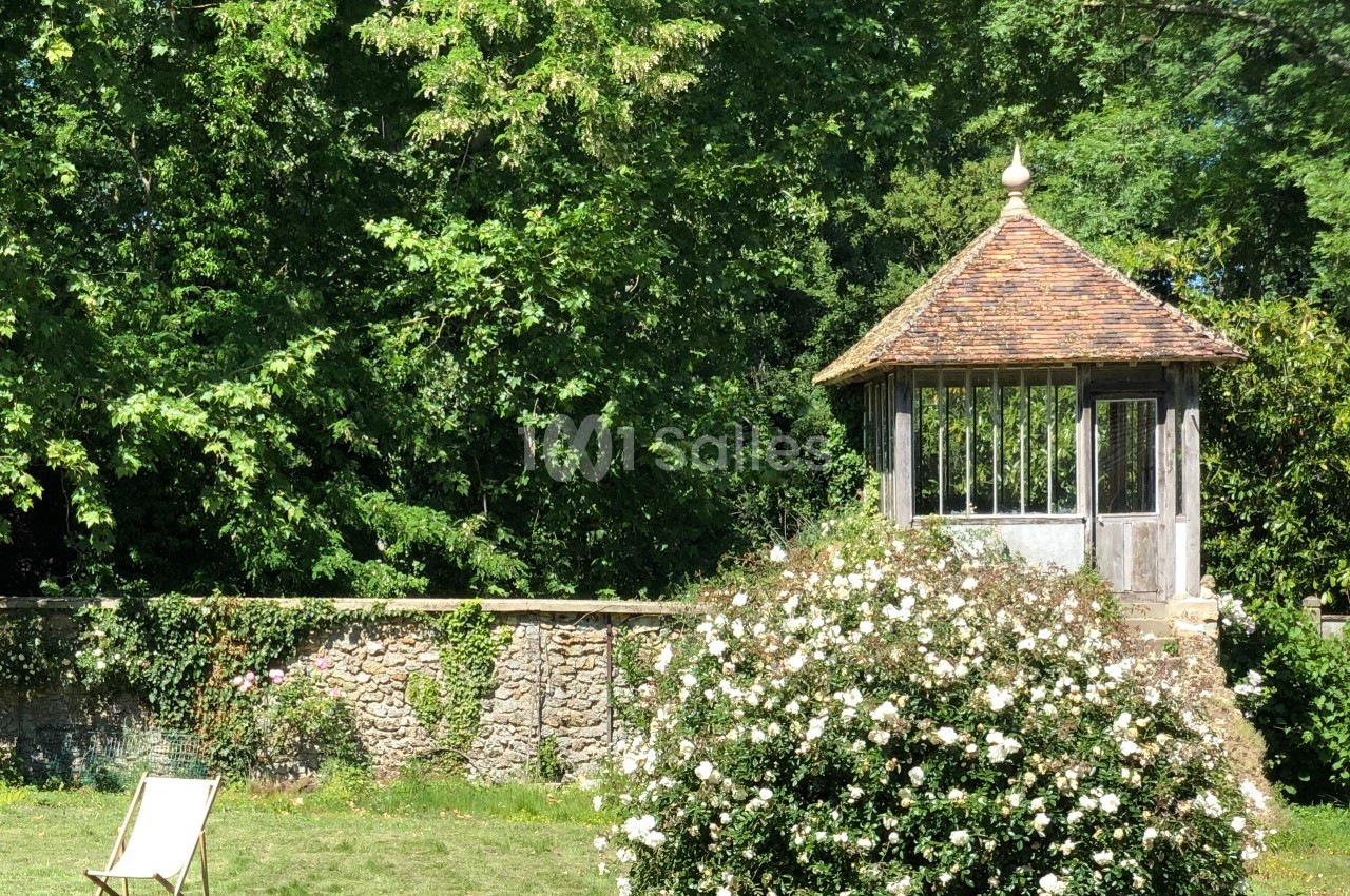 Un jardin verdoyant avec un kiosque en bois, un mur de pierre, des rosiers en fleurs et une chaise longue.
