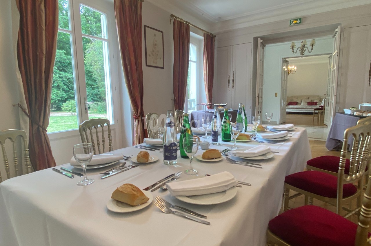 Table dressée avec nappes blanches, couverts, verres, bouteilles d'eau et pain, dans une salle lumineuse avec vue sur un…