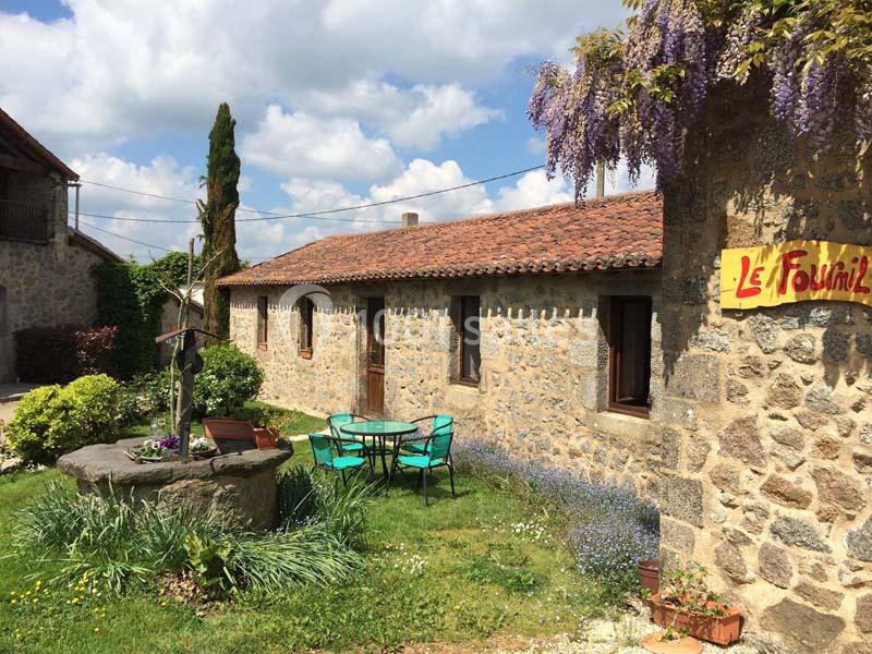 Façade en pierre d'une maison avec puits, table et chaises dans un jardin fleuri sous un ciel partiellement nuageux.