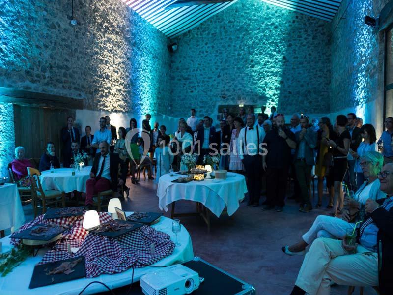 Salle en pierre éclairée en bleu, avec des invités debout et assis autour de tables décorées pour un événement.