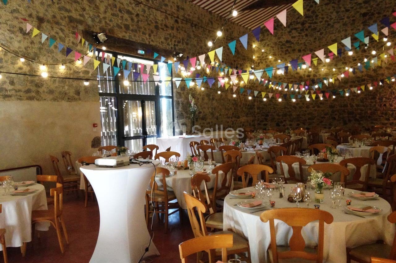 Salle décorée pour un événement avec guirlandes colorées, tables rondes dressées et murs en pierre apparente.