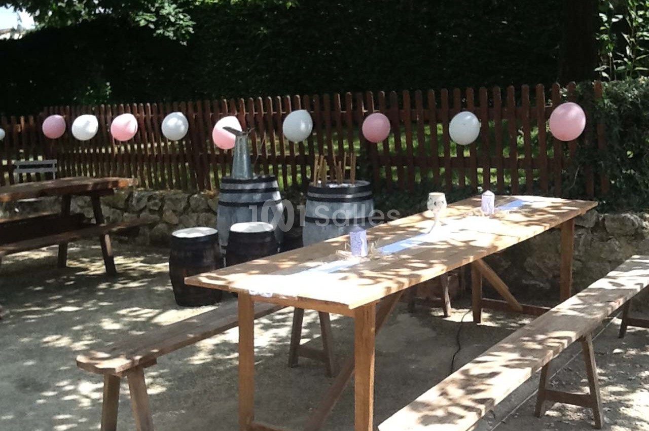Tables en bois et bancs disposés en extérieur, décorés de ballons roses et blancs près d'une clôture en bois.