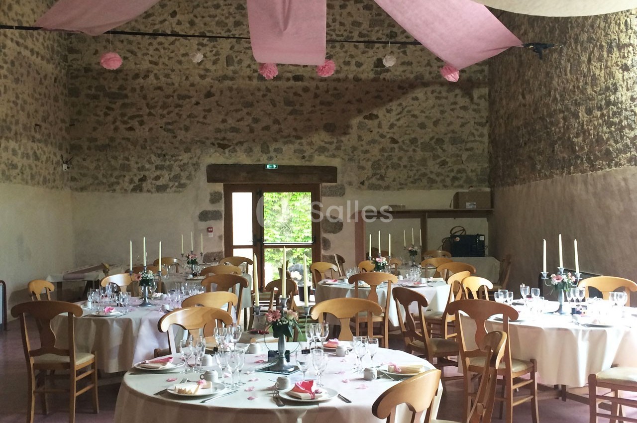 Salle de réception décorée pour un événement, avec tables rondes dressées, nappes blanches et guirlandes roses suspendues.