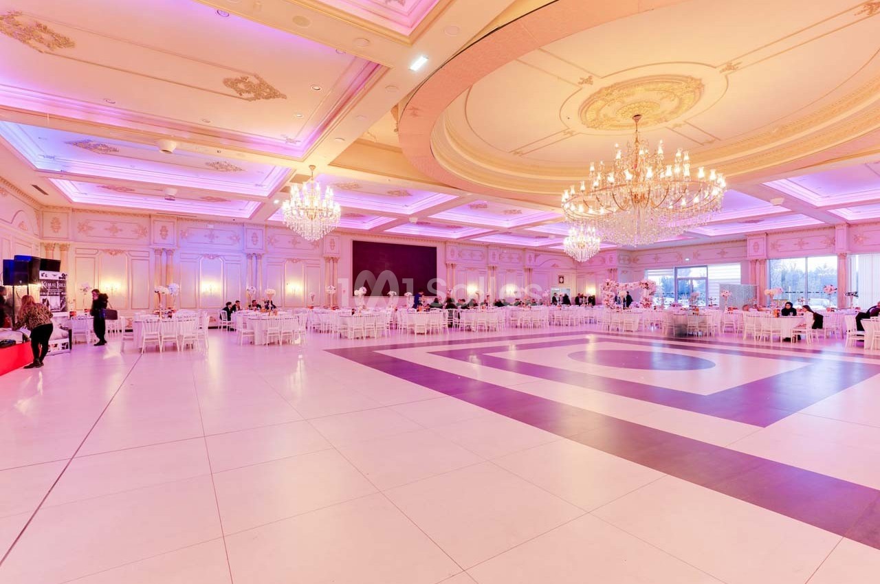 Salle de réception spacieuse avec un éclairage rose, des lustres imposants et des tables dressées pour un événement.