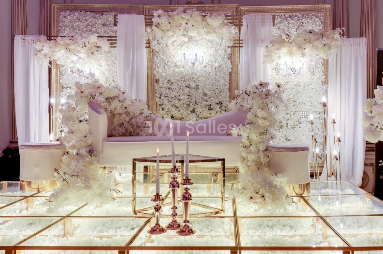 Décor élégant avec canapé blanc, fleurs blanches abondantes et chandeliers sur un sol en verre lumineux.