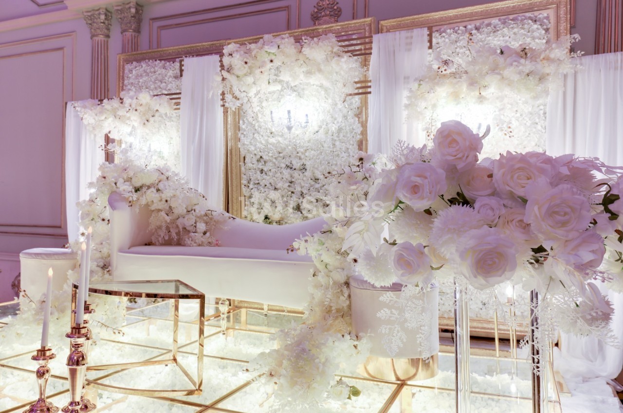 Décor de mariage élégant avec canapé blanc, fleurs blanches abondantes et bougies sur un sol en verre réfléchissant.