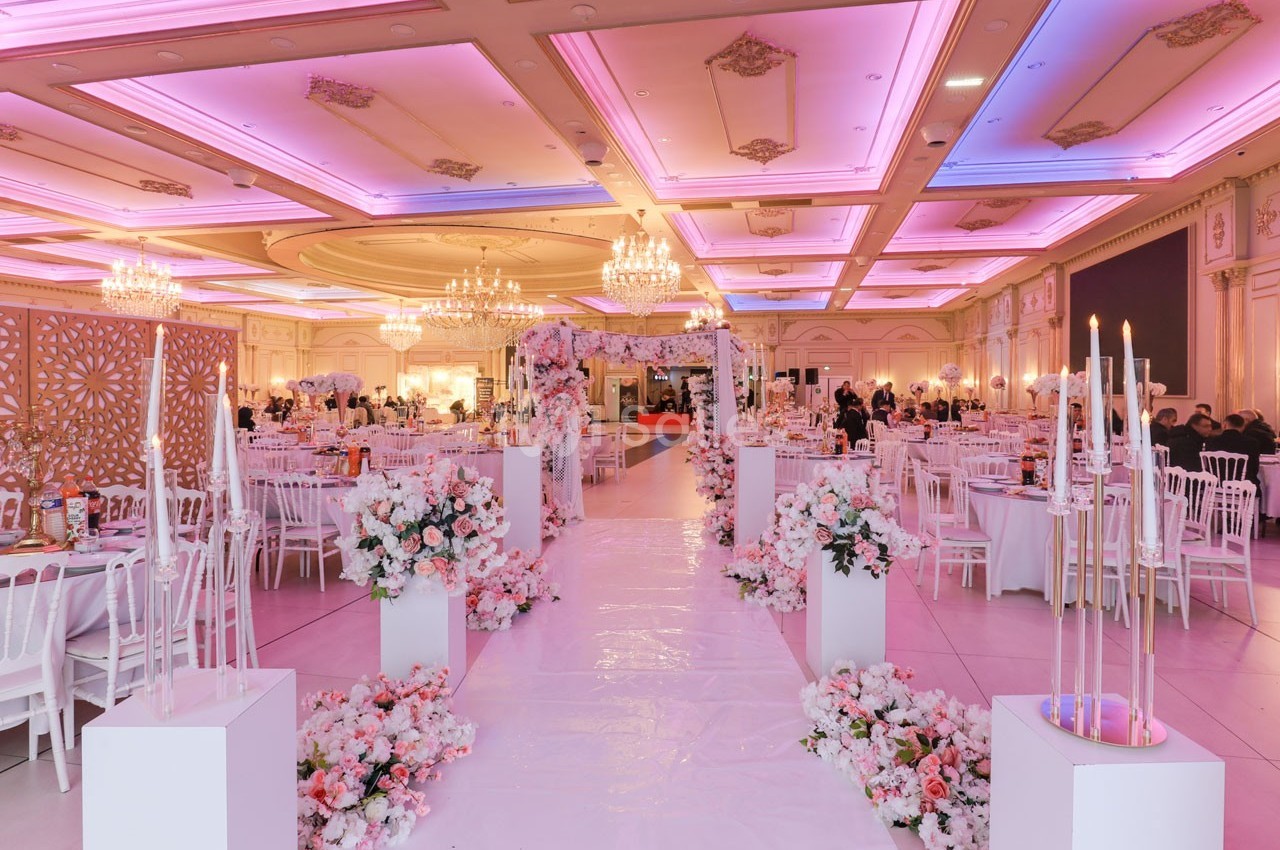 Salle de réception décorée avec des fleurs roses et blanches, chandeliers suspendus et tables élégamment dressées.