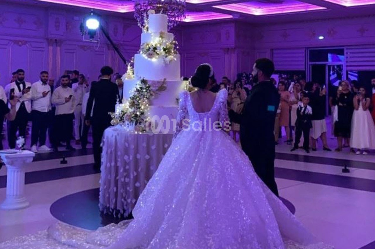 Un couple en tenue de mariage se tient devant un grand gâteau décoré, entouré d'invités dans une salle illuminée.