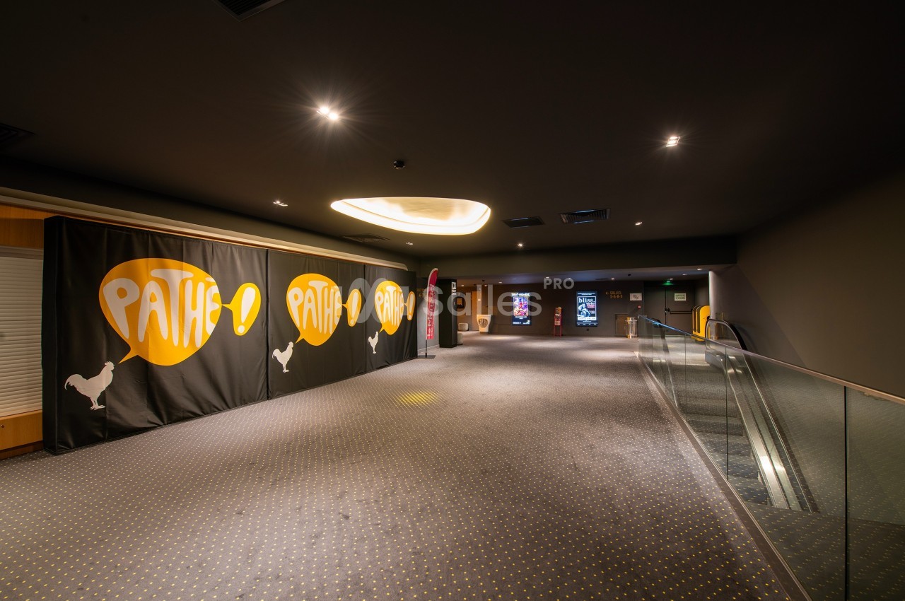 Couloir d'un cinéma avec tapis gris, panneaux lumineux au fond et escalator à droite.