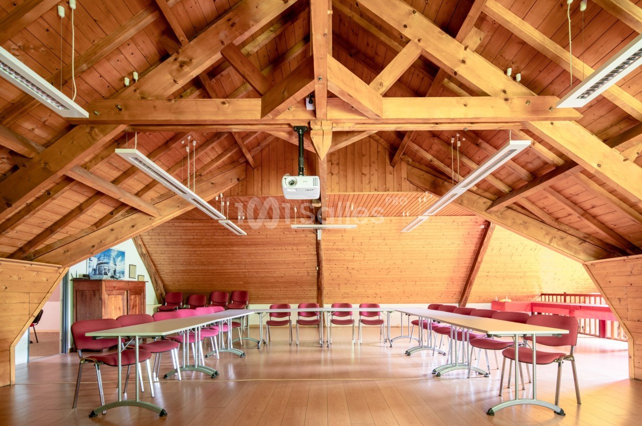 Salle de réunion lumineuse sous un toit en bois avec tables blanches, chaises rouges et projecteur suspendu.