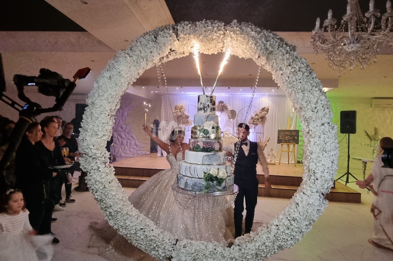 Un couple de mariés pose devant un grand gâteau entouré d'une arche florale, dans une salle de réception décorée.