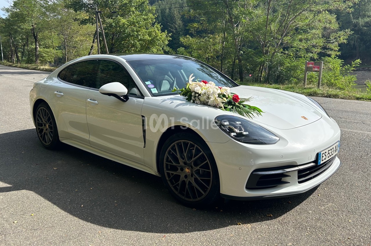 Voiture blanche décorée de fleurs blanches et vertes, stationnée sur une route bordée d'arbres.