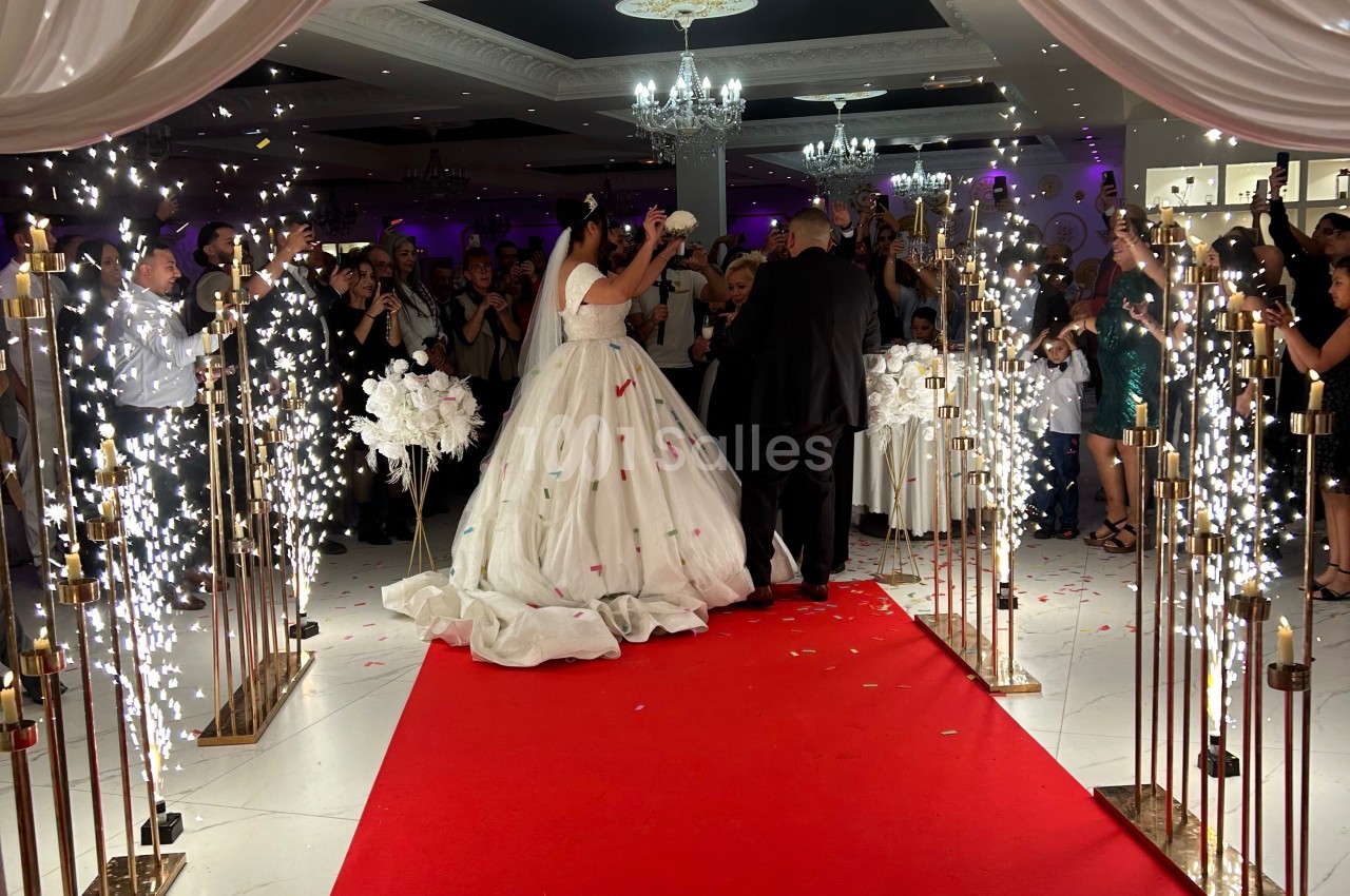 Mariés avançant sur un tapis rouge entourés d'invités, de jets d'étincelles et de décorations festives.