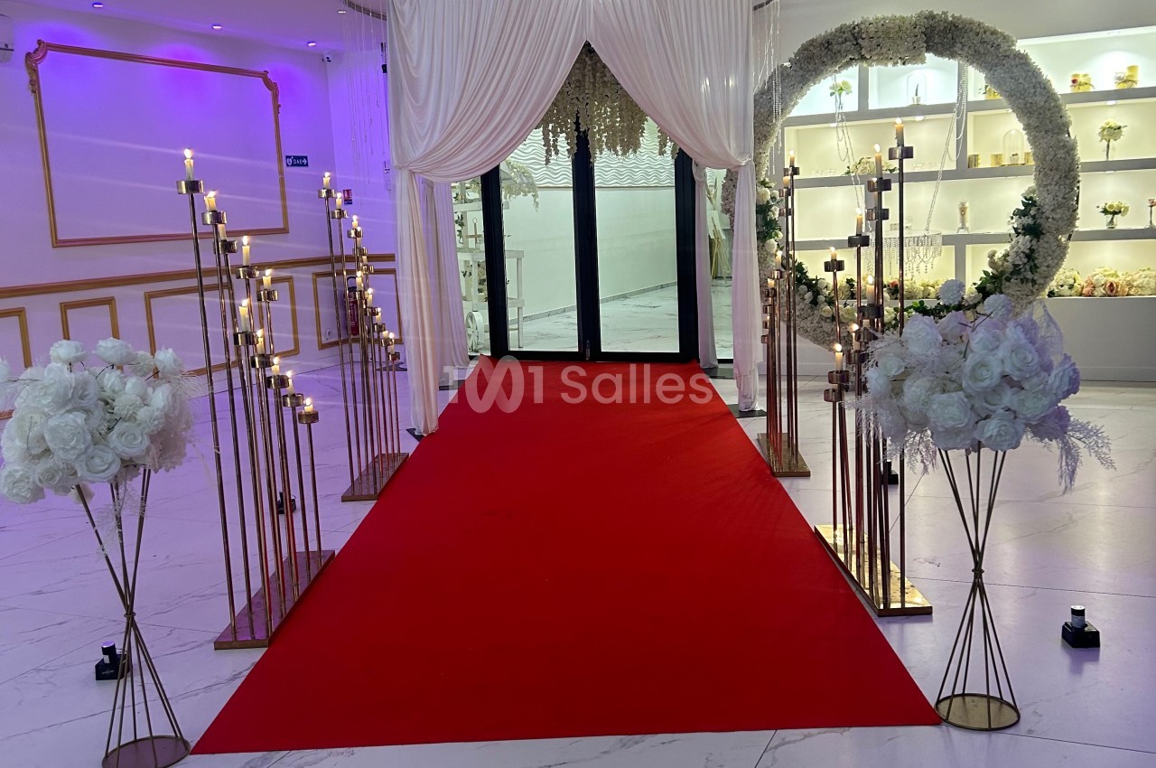 Allée décorée de chandeliers et de fleurs blanches menant à une porte, avec un tapis rouge et des rideaux blancs.