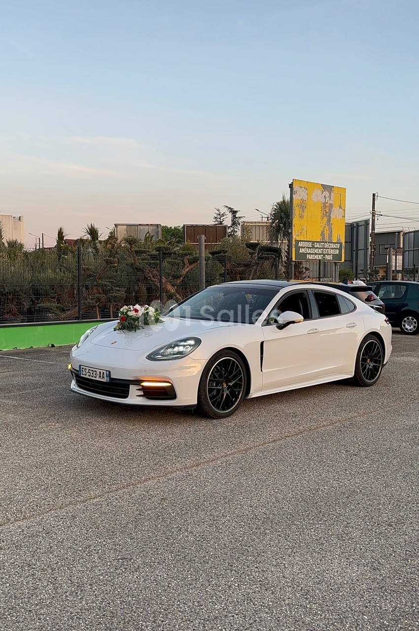 Voiture blanche décorée de fleurs à l'avant, stationnée sur une route près d'une clôture et d'un panneau publicitaire.