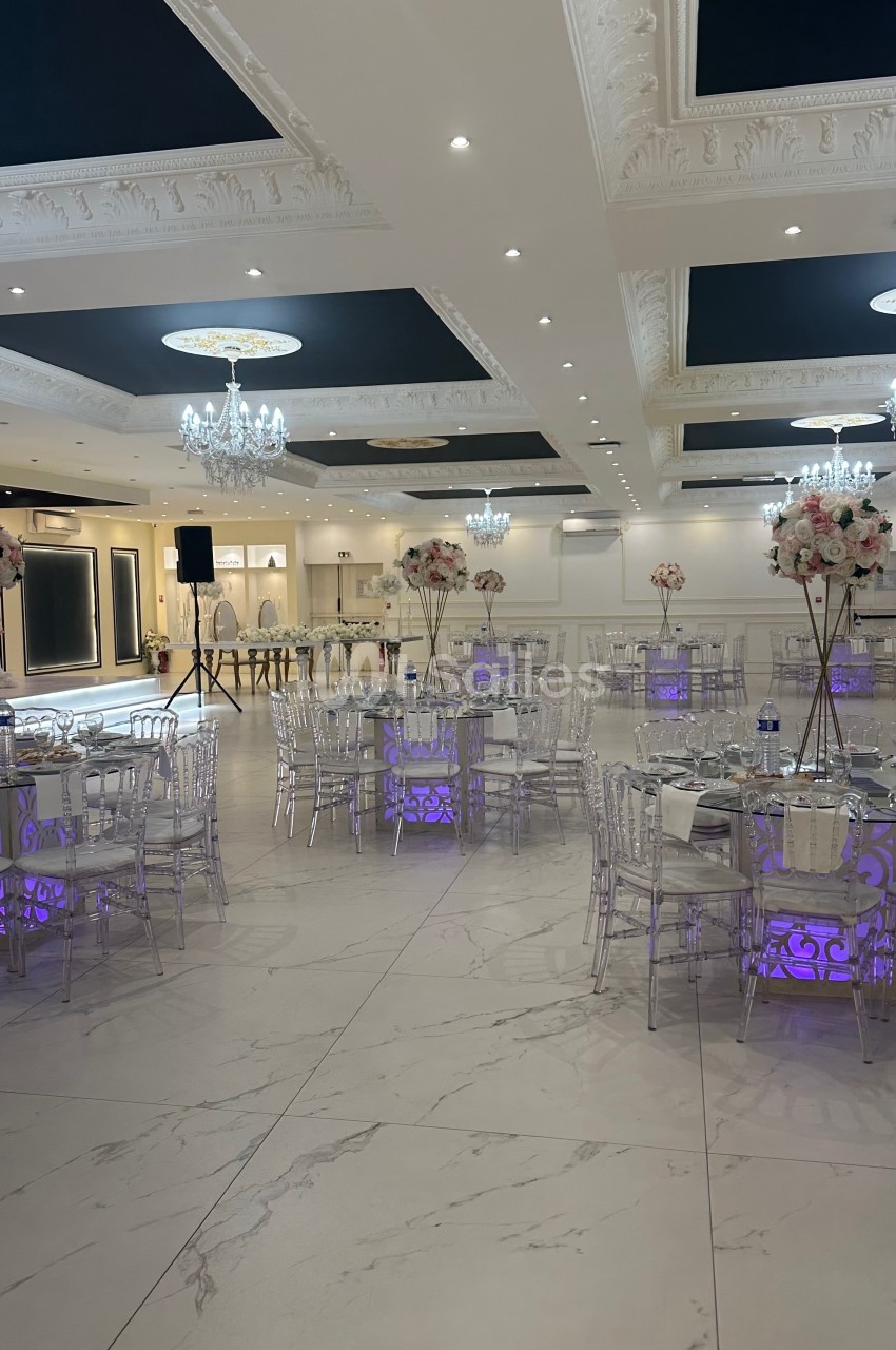 Salle de réception élégante avec tables décorées de fleurs, chaises transparentes et éclairage violet au sol.