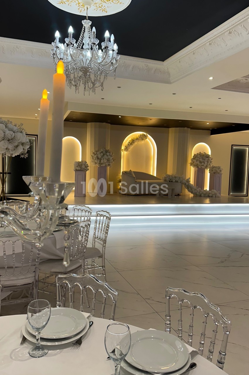 Salle de réception élégante avec table dressée au premier plan et scène décorée de fleurs en arrière-plan.