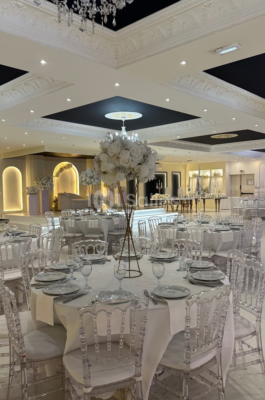 Salle de réception élégante avec tables rondes dressées, chaises transparentes et décoration florale blanche.