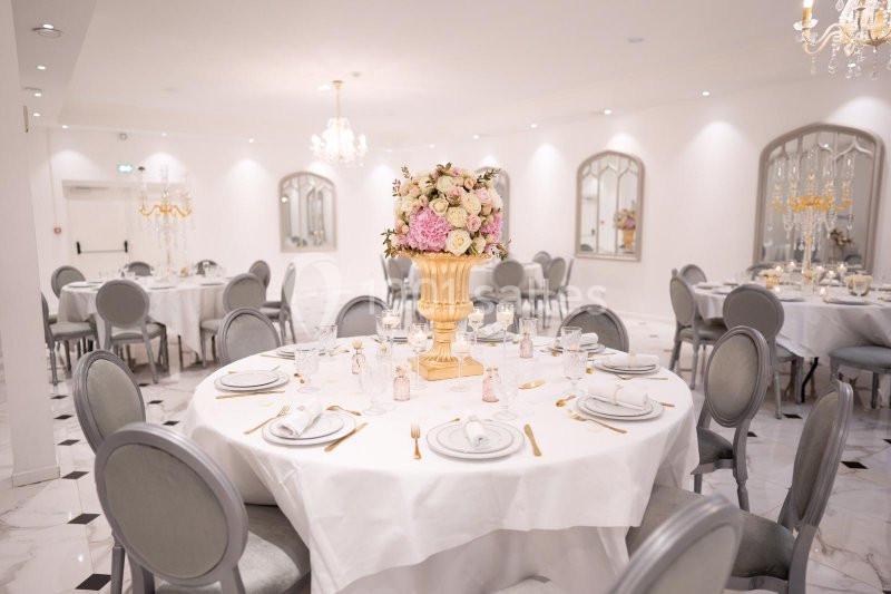Salle de réception élégante avec tables rondes dressées, chaises grises et centre de table floral au premier plan.
