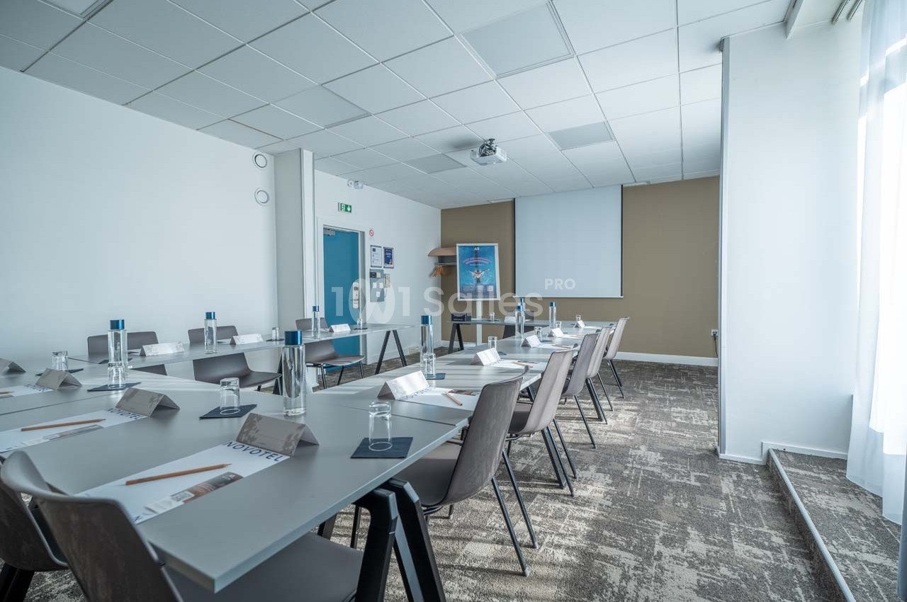 Salle de réunion lumineuse avec tables disposées en U, chaises, blocs-notes, bouteilles d'eau et écran de projection.
