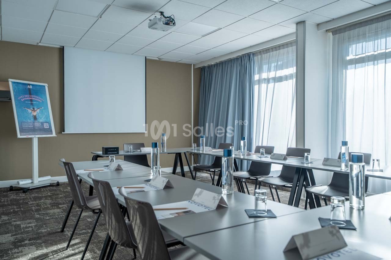 Salle de réunion lumineuse avec tables disposées en U, bouteilles d'eau, porte-noms et écran de projection.