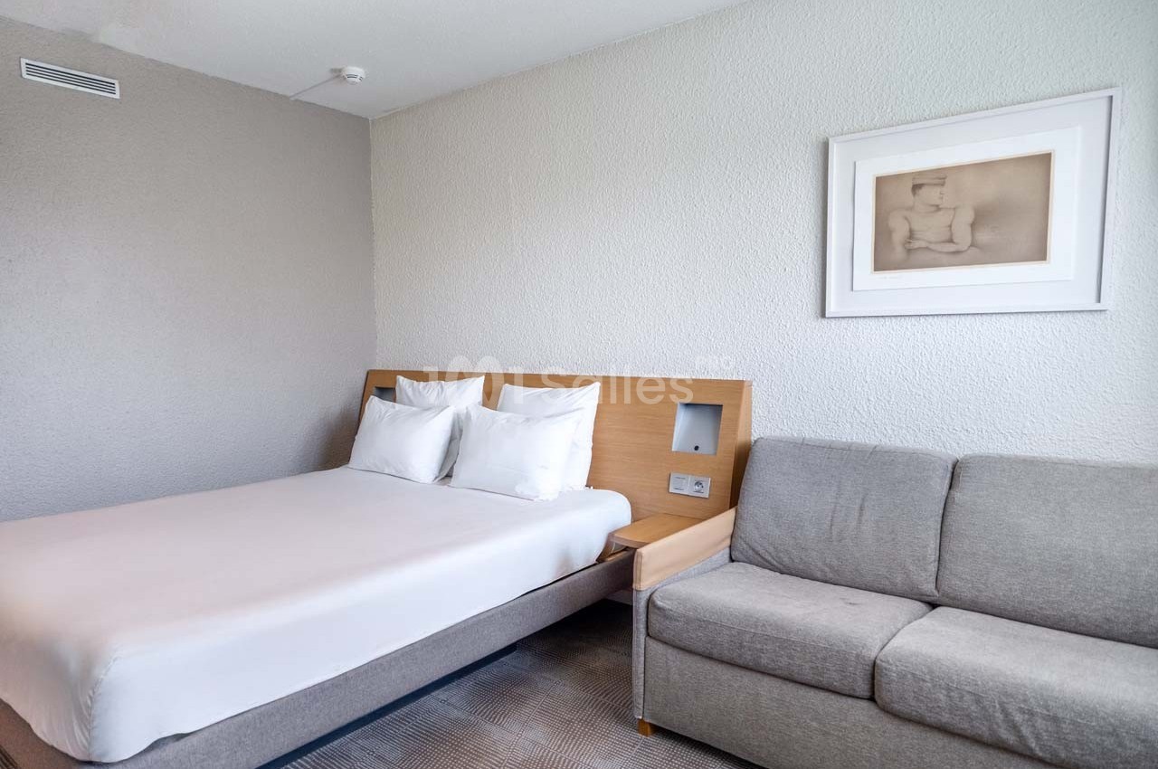 Chambre avec un lit double, un canapé gris, un mur clair et un tableau encadré.