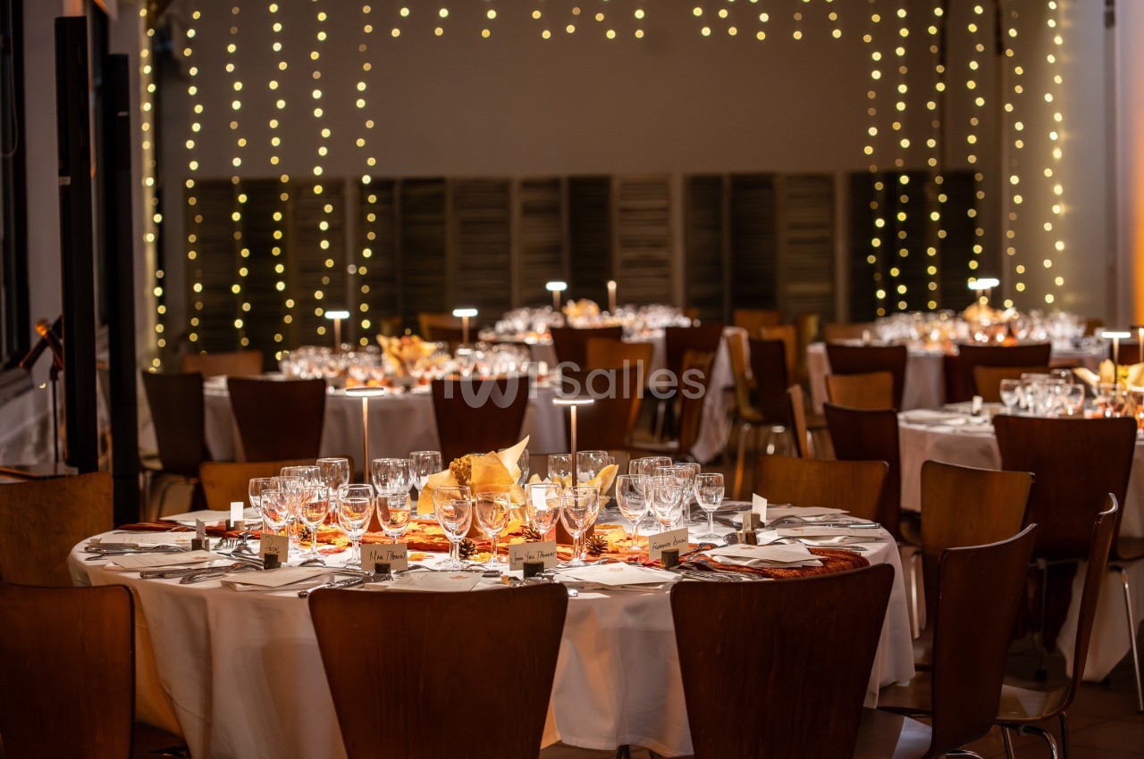 Salle de réception décorée avec des tables rondes, nappes blanches, vaisselle élégante et guirlandes lumineuses suspendues.