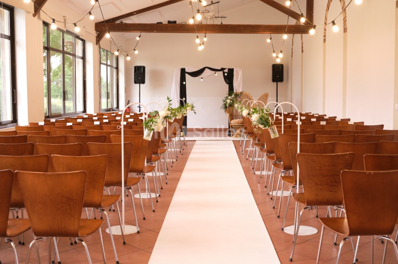 Salle décorée pour une cérémonie, avec des rangées de chaises en bois, une allée centrale et un fond orné de rideaux noirs.