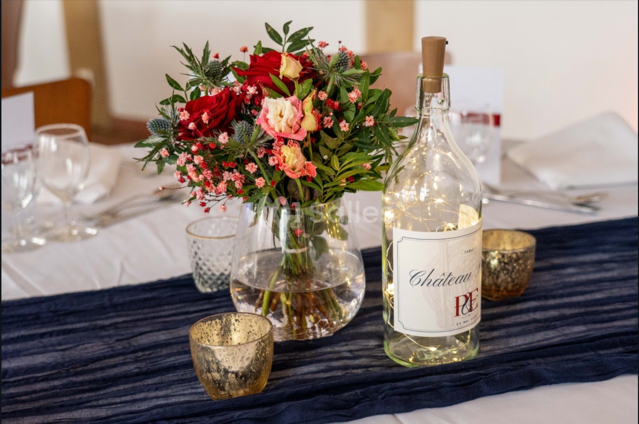 Bouquet de fleurs colorées et bouteille décorative illuminée posés sur une table avec nappe blanche et chemin bleu.