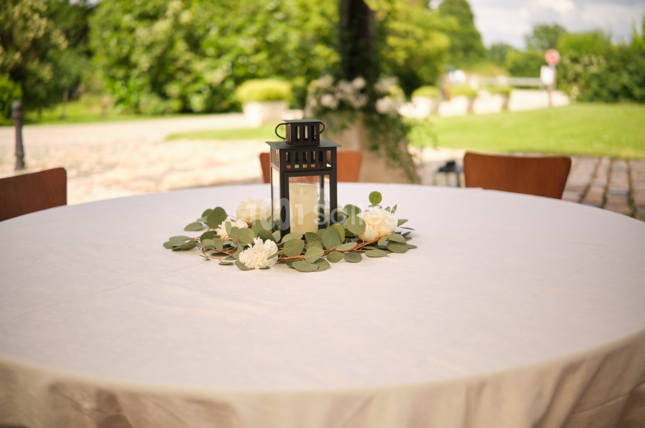 Centre de table avec une lanterne noire, des fleurs blanches et des feuilles vertes sur une nappe blanche en extérieur.