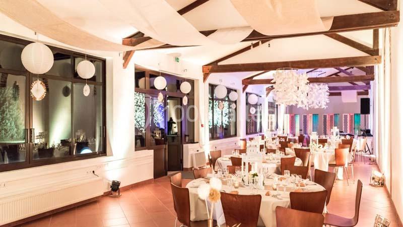 Salle de réception lumineuse avec tables dressées, décorations suspendues et grandes fenêtres donnant sur l'extérieur.
