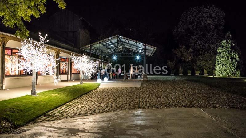 Extérieur d'un bâtiment éclairé la nuit, avec arbres lumineux, terrasse couverte et allée pavée.
